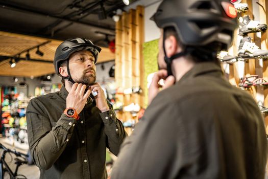 Man examining bicycle helmets in sport shop looking at herself in mirror. Trying new sports helmet in bike shop. Male putting on cycle helmet in shop. Customer in bicycle store trying on bike helmet.