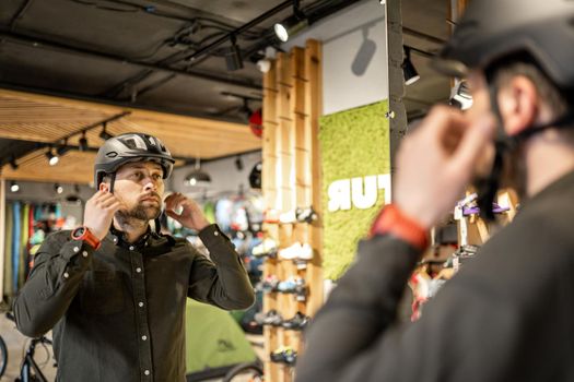 Man examining bicycle helmets in sport shop looking at herself in mirror. Trying new sports helmet in bike shop. Male putting on cycle helmet in shop. Customer in bicycle store trying on bike helmet.