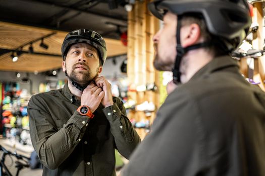Caucasian man trying on black bicycle helmet near mirror in sporting goods store. Male buyer chooses safety helmet for cycling. Shopping in bicycle store. Person adjusts his sports helmet strap.