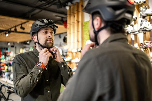 Man examining bicycle helmets in sport shop looking at herself in mirror. Trying new sports helmet in bike shop. Male putting on cycle helmet in shop. Customer in bicycle store trying on bike helmet.