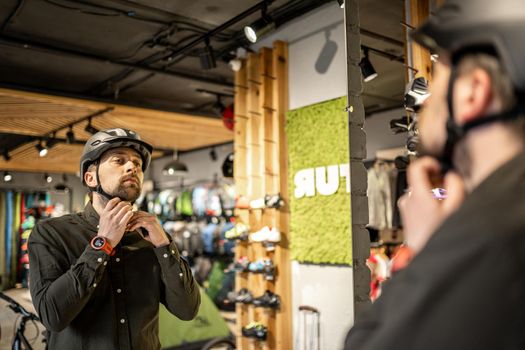 Young man came to the bicycle store. He is measuring the helmet. Male chooses helmet in sports equipment store. Purchase of new sports helmet. Customer with bicycle helmet trying on near the mirror.