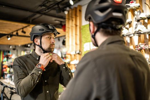 Young man came to the bicycle store. He is measuring the helmet. Male chooses helmet in sports equipment store. Purchase of new sports helmet. Customer with bicycle helmet trying on near the mirror.