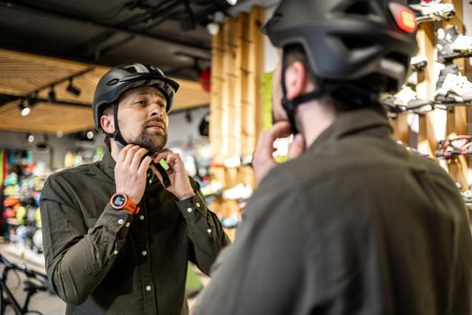 Young man came to the bicycle store. He is measuring the helmet. Male chooses helmet in sports equipment store. Purchase of new sports helmet. Customer with bicycle helmet trying on near the mirror.