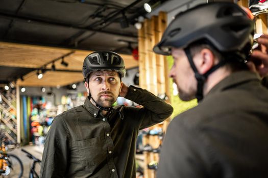Young man came to the bicycle store. He is measuring the helmet. Male chooses helmet in sports equipment store. Purchase of new sports helmet. Customer with bicycle helmet trying on near the mirror.