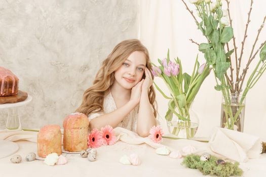A girl with long hair in a light dress is sitting at the Easter table with cakes, spring flowers and quail eggs. Happy Easter celebration