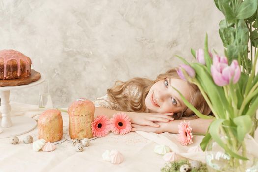 A girl with long hair in a light dress is sitting at the Easter table with cakes, spring flowers and quail eggs. Happy Easter celebration