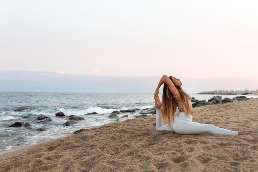 Young flexible caucasian woman practicing yoga on the beach at sunset. Copy space. Spirituality and healthy lifestyle concept.