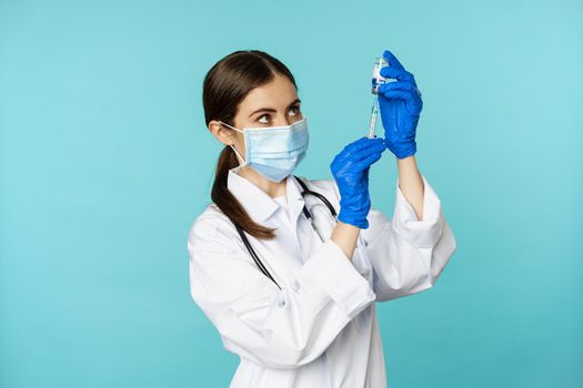 Vaccination from covid and healthcare concept. Young woman doctor, nurse in face mask and gloves, using syringe to do vaccine shot, standing over torquoise background.