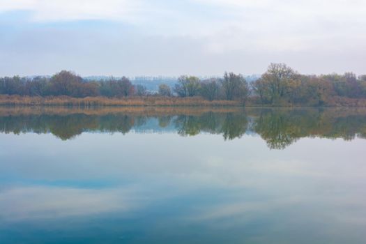 Cold foggy river in the morning with overgrown banks and displaying clouds in the water. Late autumn.