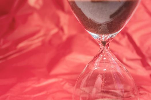 Hourglass on a red wavy silk background. Luxury and time concept concept. Nothing is eternal. The sand passes through the glass bulbs of the watch. Place for your text.