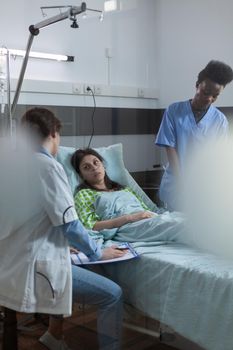 Doctor holding clipboard with lab results sitting down talking with recovering patient about treatment options. African american nurse giving medical care to ill woman in hospital ward.