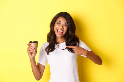 Image of cheerful african-american woman, recommending good coffee from cafe, pointing at cup, standing over yellow background.