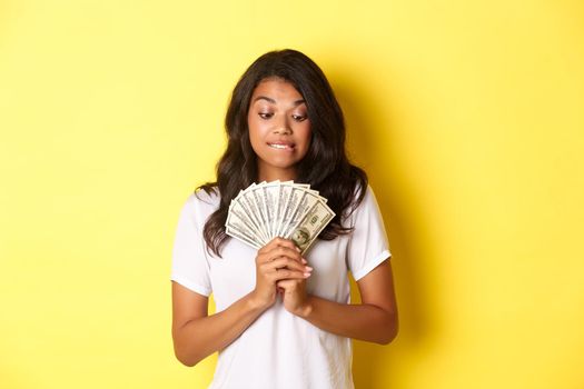 Portrait of attractive african-american woman, looking tempted at money, wanting to buy something, standing over yellow background.