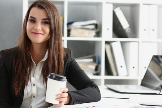 Portrait of attractive worker in suit holding cup of coffee, posing on working place in office. Break for lunch, enjoy drink, pause. Chill, relax concept