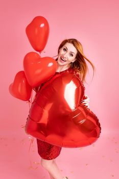 Beautiful redhead girl with red heart baloon posing. Happy Valentine's Day concept. Studio photo of beautiful ginger girl dancing on pink background.