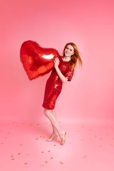 Beautiful redhead girl with red heart baloon posing. Happy Valentine's Day concept. Studio photo of beautiful ginger girl dancing on pink background.