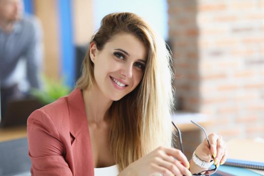 Portrait of attractive blonde lady sit in personal cabinet holding glasses. Smiling employee in stylish pink suit work in big corporation. Business concept