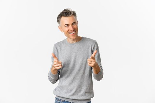 Cheeky middle-aged man with grey short hairstyle, pointing fingers at camera and smiling, praising good work or congratulating, standing over white background.