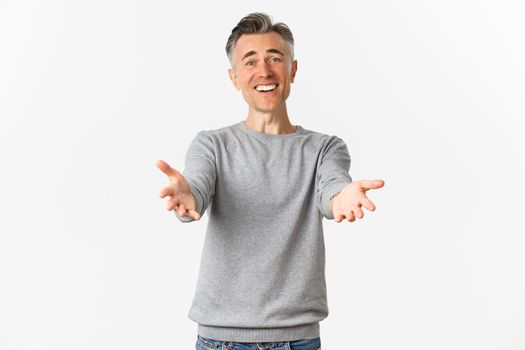 Image of happy and pleased middle-aged man with gray hair, reaching hands forward to hug or welcome someone, smiling relieved, standing over white background.
