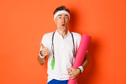 Concept of workout, gym and lifestyle. Close-up of happy and satisfied middle-aged athlete, showing thumb-up, like training session, holding yoga mat and skipping rope for fitness.