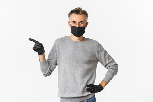 Concept of coronavirus, lifestyle and quarantine. Portrait of serious middle-aged guy in medical mask and gloves, pointing finger left at copy space, standing over white background.