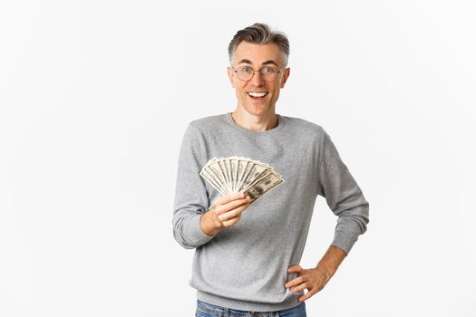 Portrait of amazed middle-aged man in glasses and gray sweater, holding money and smiling cheerful, winning lottery, standing over white background.