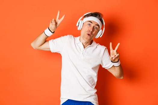 Portrait of carefree middle-aged male athlete, listening music in headphones during fitness training, showing peace signs, standing over orange background.