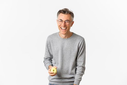 Image of happy and healthy middle-aged man, holding green apple, laughing and smiling carefree, standing over white background.