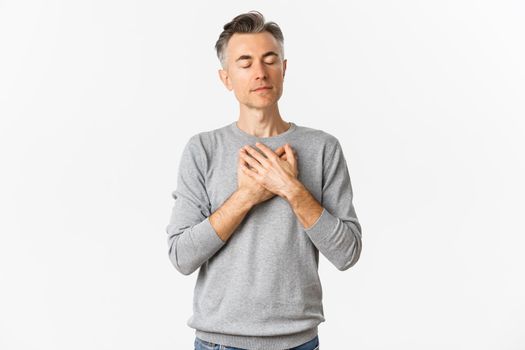 Image of handsome middle-aged male model with gray hair, holding hands on heart and thinking about something with eyes closed, daydreaming, standing over white background.