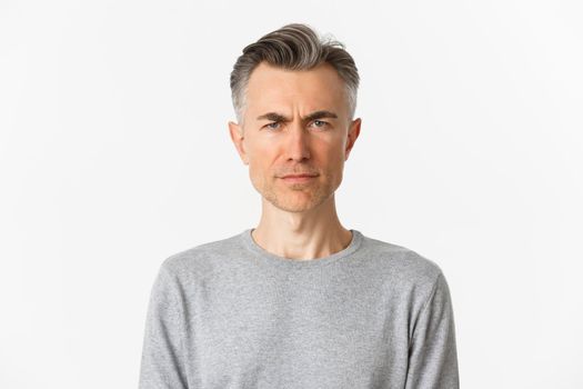 Close-up of skeptical middle-aged man, frowning and looking doubtful at camera, standing over white background in gray sweater.