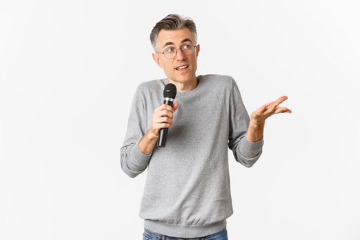 Portrait of handsome middle-aged man in glasses and gray sweater, making speech, talking with microphone and explaining something, standing over white background.