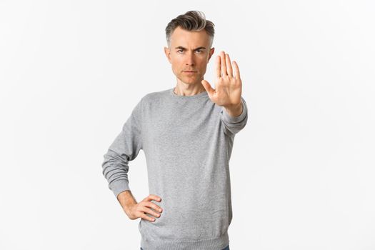 Image of serious middle-aged man extend one hand to stop you, prohibit something bad, frowning and looking with disapproval, standing over white background.