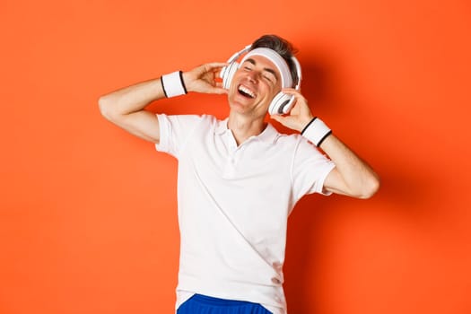 Portrait of carefree middle-aged male athlete, listening music in headphones during fitness training, standing over orange background.