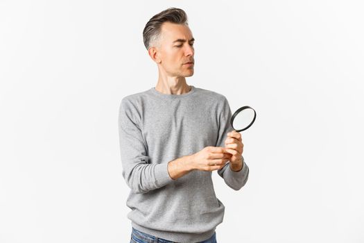 Portrait of thoughtful middle-aged man looking through magnifying glass, searching for something or reading, standing over white background.