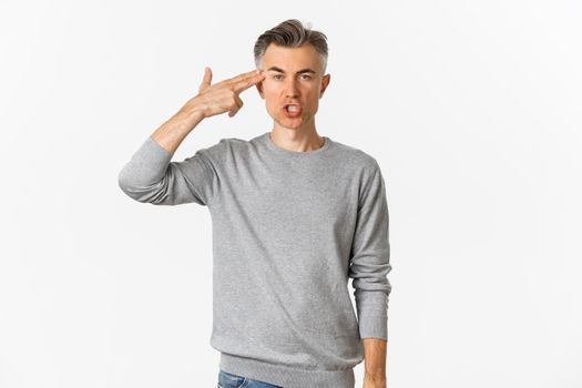 Image of annoyed and fed up middle-aged man showing finger gun sign near head, shooting himself with pissed-off face, standing tired over white background.