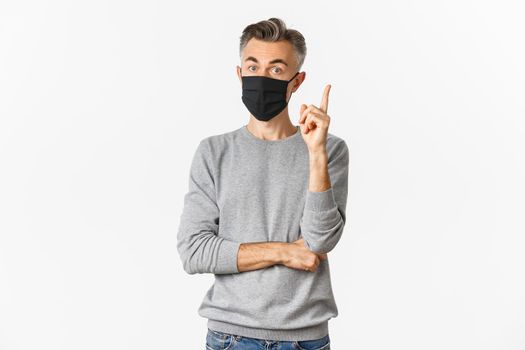 Covid-19, pandemic and social distancing concept. Portrait of handsome middle-aged caucasian man, wearing medical mask, having suggestion or idea, raising finger in eureka sign, white background.