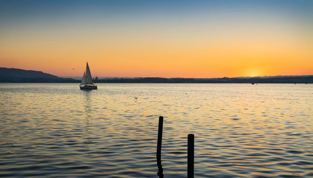 Landscape at sunset on Lake Zugersee in the Swiss town of Zug, with a sailboat crossing the horizon and the warm tones reflecting on the water.