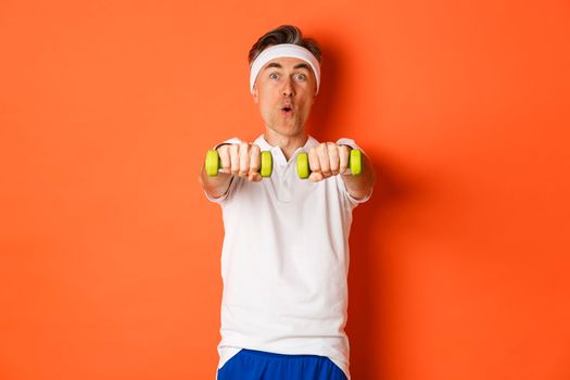 Image of active middle-aged fitness guy, doing sport exercises with dumbbells and smiling excited, standing over orange background.