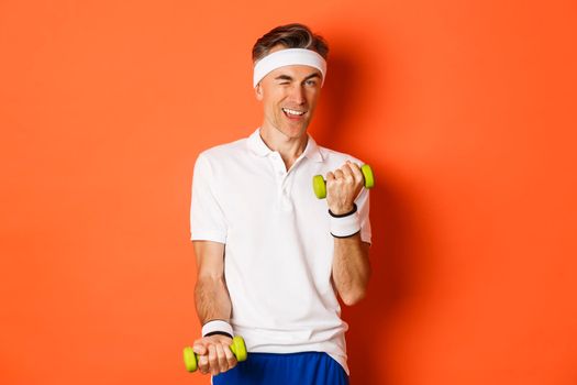 Portrait of handsome cheeky, middle-aged male athlete doing sports, exercise with dumbbells and winking at camera, standing over orange background.