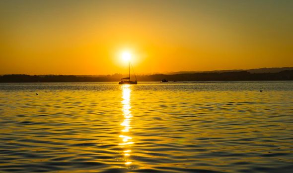 Sunset landscape reflecting on a lake with the silhouette of a sailboat on the horizon silhouetted against the sunlight.