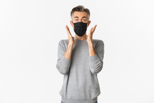 Covid-19, pandemic and social distancing concept. Image of surprised middle-aged man in black medical mask looking at camera, stare amazed at something awesome, white background.