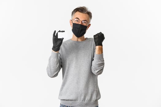 Concept of covid-19, social distancing and lifestyle. Image of attractive middle-aged man in medical mask, gloves and glasses, showing credit card and rejoicing, standing over white background.