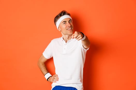 Concept of sport, fitness and lifestyle. Portrait of confident, healthy middle-aged sportsman, pointing finger at camera and smiling, need you, standing over orange background.