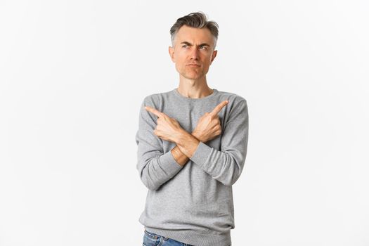 Image of indecisive middle-aged man making choice, pointing fingers sideways and frowning doubtful, standing unsure over white background.