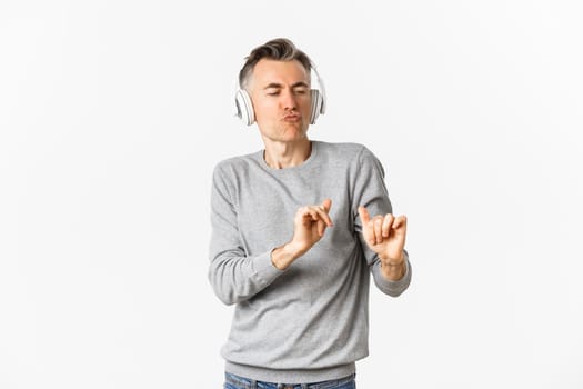 Portrait of handsome, modern middle-aged man, listening music in headphones and dancing, standing over white background.