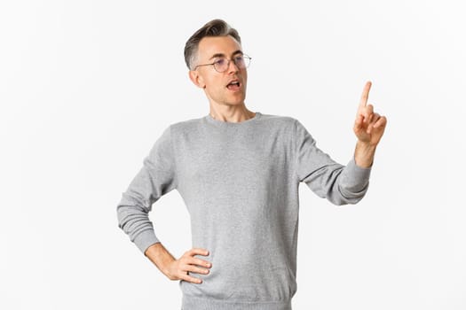 Image of confident middle-aged businessman in grey sweater and glasses, pointing finger and making speech, giving tasks to employees, standing over white background.
