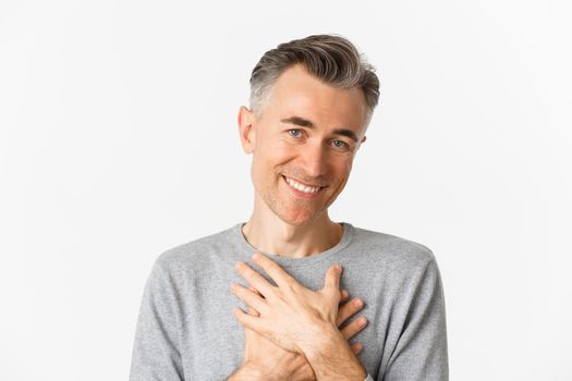 Portrait of thankful middle-aged man, holding hands on heart and smiling, feeling touched and grateful, standing over white background.