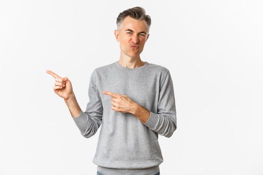 Portrait of skeptical and disappointed middle-aged man, grimacing and pointing fingers left at bad promo, standing over white background.