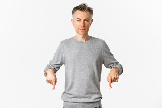 Portrait of successful middle-aged man in grey sweater, pointing fingers down and looking confident at camera, showing logo, standing over white background.