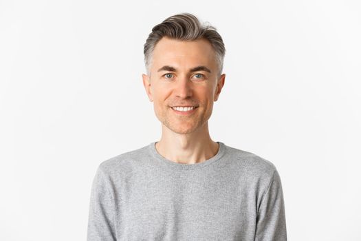 Close-up of handsome middle-aged man in gray sweater, looking happy and hopeful, smiling at camera, standing over white background.